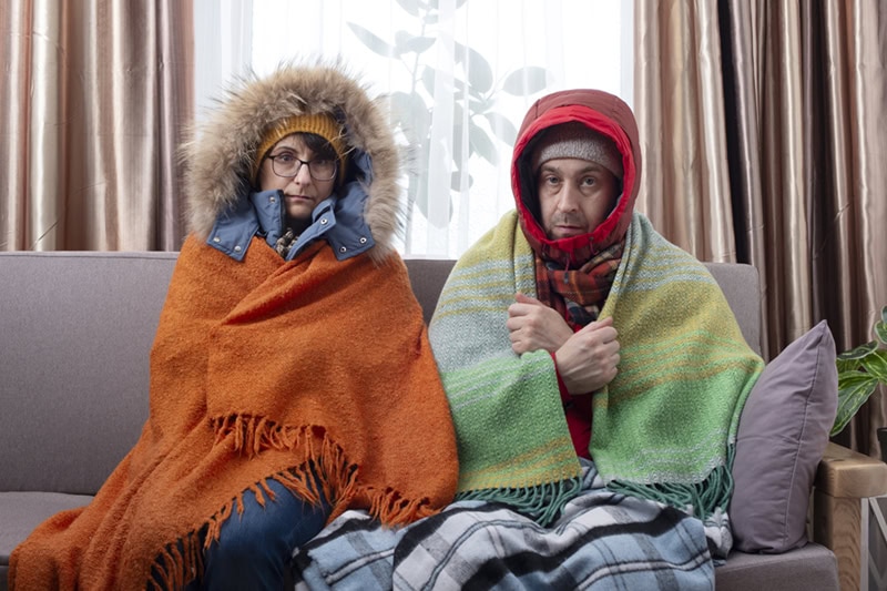 A shivering man and woman sit bundled up on a living room sofa, wearing heavy hooded coats, hats, and warm blankets wrapped around themselves to keep warm indoors.