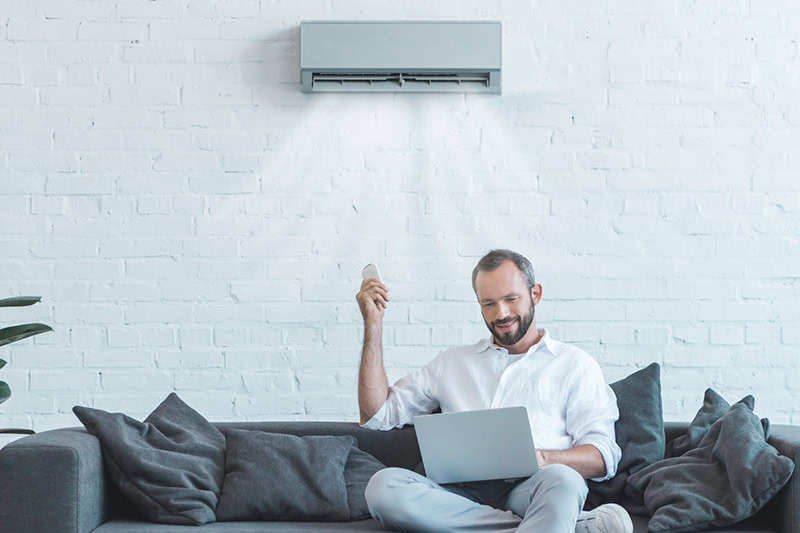 A man sitting on his couch looking at his laptop. He is holding a remote which turns on his ductless mini split system.